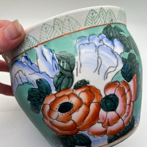 Könnte beinhalten: Eine Keramikschale mit handgemaltem Blumendesign. Die Schale zeigt orange, rosa und blaue Blumen mit grünen Blättern auf einem türkisfarbenen Hintergrund. Der Rand hat einen weißen Rand mit grünen Blättermustern.