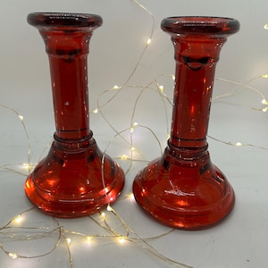 Pode incluir: Dois castiçais de vidro vermelho com base alargada e topo largo. Os castiçais são de cor vermelha profunda e têm um design clássico. O fundo inclui uma sequência de pequenas luzes brancas quentes.