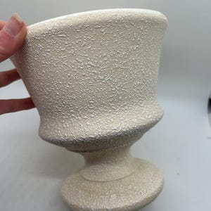 May include: Off-white textured urn-shaped vase with a pedestal base. The vase has a wide, flared rim and a textured surface. The neutral color and classic shape make it suitable for various decorative purposes.