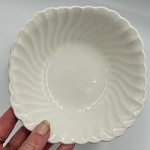 May include: A white ceramic bowl featuring a scalloped edge and a subtle, swirling ribbed pattern that radiates from the center toward the rim. The bowl has a glossy finish and a circular shape with a slightly deep basin for serving food items.
