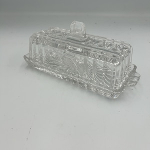 May include: A clear glass butter dish with a rectangular base and a matching lid. The lid features a central handle and intricate geometric starburst patterns etched into the glass. The base has scalloped edges and a textured, faceted design.