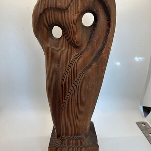 Pode incluir: Uma escultura de coruja de madeira com rosto e olhos esculpidos. A coruja está em pé sobre uma base quadrada. A escultura é feita de madeira marrom escura com grão natural.