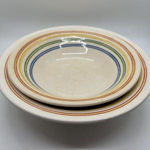 Pode incluir: Duas tigelas de cerâmica branco-sujo, uma dentro da outra. Cada tigela apresenta uma série de anéis coloridos em laranja, amarelo, verde, azul e castanho. A tigela externa tem uma borda mais larga com uma única faixa laranja.