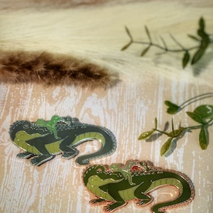 May include: Two green enamel pins of a lizard with smaller lizards on its back. The lizard on the left is on a silver base and the lizard on the right is on a copper base.