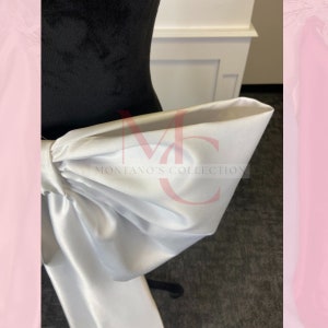 May include: A white satin bow tied around a black dress form. The bow is large and has a lot of volume. The text "MONTANO'S COLLECTION" is visible on the bow.