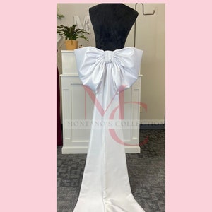 May include: White satin chair sash with a large, white bow. The sash is tied around the back of the chair and has a long, flowing tail.