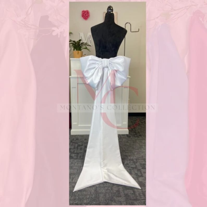 May include: A white satin bow with a long tail, attached to a white fabric backdrop. The bow is large and dramatic, with a center loop and two long tails.
