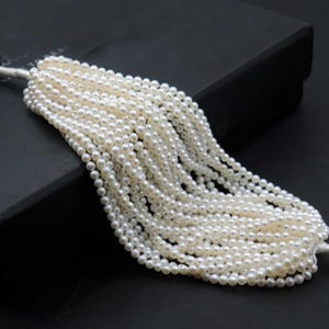 May include: Strands of small, round, white pearls cascade from a central point. The pearls are strung together, creating a flowing, elegant display. The background is a dark, solid color, which contrasts with the bright pearls.