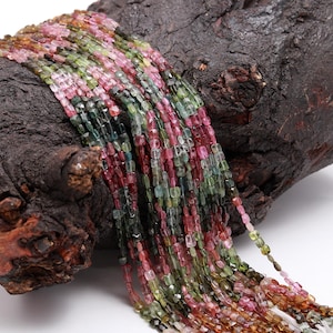 May include: A strand of multi-colored tourmaline beads, featuring shades of green, pink, and brown. The beads are faceted and have a square shape. The beads are strung on a clear thread and are displayed on a piece of wood.