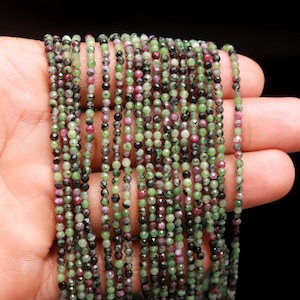 Puede incluir: Primer plano de múltiples hebras de cuentas de piedras preciosas. Las cuentas son pequeñas, facetadas y en tonos verdes, rojos y negros. Las cuentas están ensartadas, creando una textura delicada. Las cuentas se sostienen en una mano sobre un fondo negro.