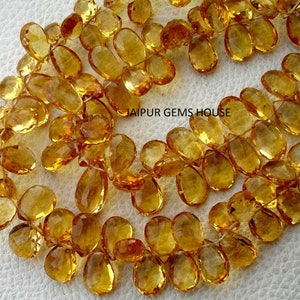 Può includere: Una fila di pietre preziose sfaccettate, a forma di goccia, di colore giallo dorato. Le pietre sono infilate su un filo trasparente e sono lunghe circa 10 mm.  JAIPUR GEMS HOUSE è stampato sull'immagine.