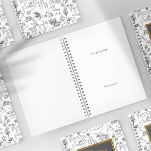 Pu&ograve; includere: Ricettario bianco a spirale con un'etichetta nera e dorata con la scritta "Ricette". La copertina presenta un'illustrazione botanica in bianco e nero. La pagina aperta riporta il testo "Le nostre ricette di famiglia" e "Questo libro appartiene a".