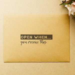 Può includere: Una busta marrone con il testo "OPEN WHEN... you receive this" stampato su di essa.