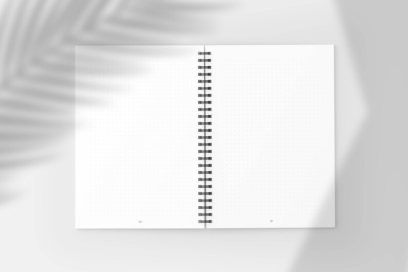 Pu&ograve; includere: Quaderno aperto a spirale con pagine bianche a puntini. Il quaderno &egrave; aperto, rivelando due pagine affiancate con una griglia di piccoli punti. La rilegatura &egrave; nera e le pagine sono bianche. Un'ombra di foglia &egrave; proiettata.