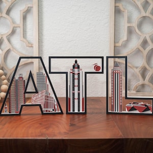 May include: Black framed wooden letters spelling "ATL" with city skyline cutouts. The "A" features red and gray buildings, the "T" a tall skyscraper, and the "L" a red and gray building. A peach graphic is above the "T".