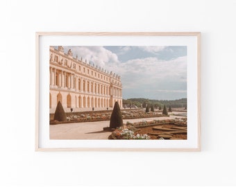 May include: Framed print featuring a pastel-toned photograph of a grand palace with a long facade, columns, and a manicured garden. The image includes cone-shaped trees, flower beds, and a blue sky with clouds. The print is in a light wood frame.