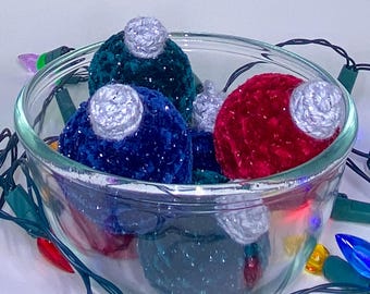 May include: A clear glass bowl filled with plush, colorful Christmas ornaments. The ornaments are in shades of red, blue, and green, with silver tops. A string of colorful Christmas lights surrounds the bowl, adding a festive touch.
