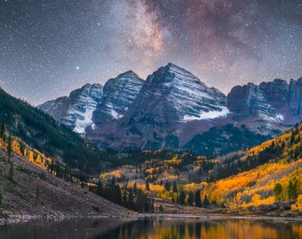 Puede incluir: Una cordillera con picos cubiertos de nieve bajo un cielo nocturno lleno de estrellas y la Vía Láctea. Las montañas están rodeadas de un bosque de árboles con hojas amarillas y naranjas. Un lago refleja el cielo y las montañas.