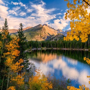 May include: A scenic view of a mountain lake with a mountain range in the background. The lake is surrounded by trees, some of which have yellow leaves. The sky is blue with white clouds.