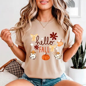 Puede incluir: Camiseta beige con las palabras "hello FALL" en marrón y naranja. La camiseta presenta ilustraciones de una calabaza, una taza de café y dos figuras fantasmales con sombreros de vaquero. La camiseta tiene las mangas enrolladas.