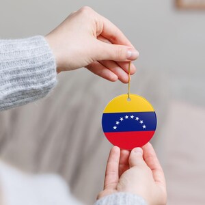 Puede incluir: Adorno redondo con la bandera de Venezuela. El diseño incluye una parte superior amarilla, un centro azul con estrellas blancas y una parte inferior roja. Colgando de un cordón dorado.
