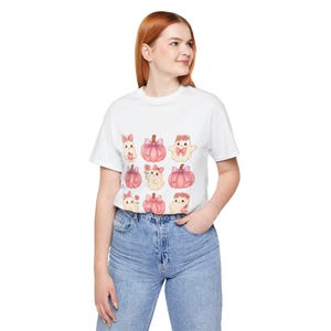 Puede incluir: Camiseta blanca con un patrón repetido de calabazas y fantasmas de dibujos animados, adornados con lazos rosas y detalles florales. El diseño utiliza tonos suaves de rosa, melocotón y blanco. La camiseta la lleva una persona con vaqueros azules.