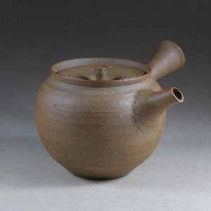 May include: A brown ceramic teapot with a rounded body, a side-mounted handle, a short spout, and a flat lid featuring a small central knob. The surface has a matte, textured finish with subtle variations in the earthy brown clay tone.