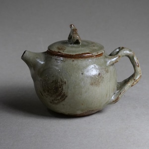 Può includere: Piccola teiera artigianale in ceramica, di colore verde tenue con accenti marroni e superficie strutturata. Il manico ha un design attorcigliato e il coperchio presenta un piccolo pomello decorativo. Per servire il tè.