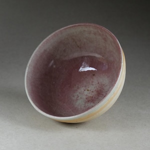 Può includere: Una ciotola in ceramica dalla forma arrotondata. L'interno è di un colore bordeaux intenso con sottili macchioline, mentre l'esterno presenta una fascia color crema e giallo chiaro. La ciotola ha una finitura lucida ed è posta su uno sfondo neutro.