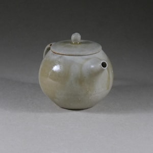 Op de afbeelding: Een ronde keramische theepot in een gedempte groene tint, met een deksel en een klein handvat. De theepot heeft een glanzende afwerking en een kleine, ronde tuit. Het deksel is bekroond met een kleine knop.