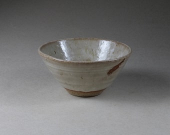 May include: A handmade ceramic bowl with a wide, shallow design. The bowl has a creamy white glaze with subtle variations and a brown rim and base. A brown streak accents the interior.