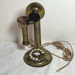 May include: Antique brass candlestick telephone with a rotary dial. The phone features a black earpiece, a wooden mouthpiece, and a coiled cord. The base has a circular dial with white numbers and letters. A vintage communication device.