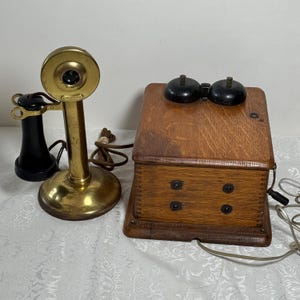 Puede incluir: Un teléfono antiguo que presenta un transmisor de latón tipo candelabro con un auricular negro y una base rectangular de madera. La unidad de madera incluye dos campanas de metal negro en la parte superior y una manivela lateral, unida por un cable.