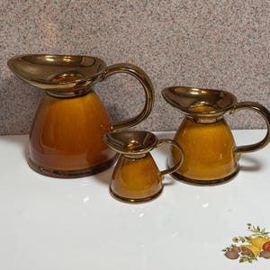 Può includere: Tre brocche in ceramica di diverse dimensioni. Le brocche hanno una glassa marrone con bordo e manico dorati. La brocca più grande è alta circa 18 cm, la brocca media 13 cm e la più piccola 8 cm.