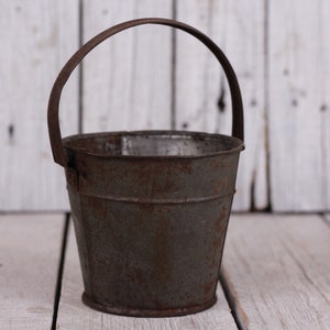 Könnte beinhalten: Ein kleiner, rostiger Metalleimer mit Henkel. Der Eimer ist leer und hat ein abgenutztes, altes Aussehen.