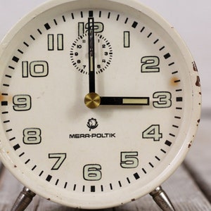 May include: White vintage alarm clock with black numbers and hands. The clock face reads "MERA-POLITIK".