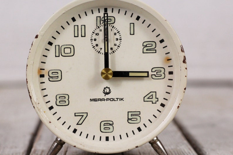 May include: White vintage alarm clock with black numbers and hands. The clock face reads "MERA-POLITIK".