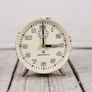 May include: White vintage alarm clock with black numbers and hands. The clock face has the text "Mera-Poltik" printed on it.