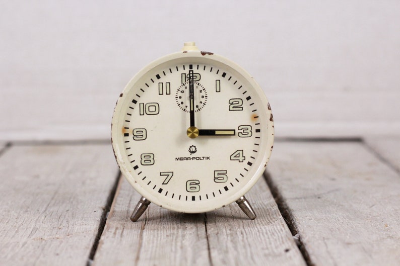 May include: White vintage alarm clock with black numbers and hands. The clock face has the text "Mera-Poltik" printed on it.