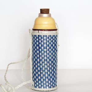 Può includere: Una borraccia vintage con un motivo a quadri blu e bianchi. La bottiglia ha una parte superiore color crema e un tappo in sughero. Un cinturino bianco intrecciato è attaccato per il trasporto. La bottiglia è alta circa 25 cm.