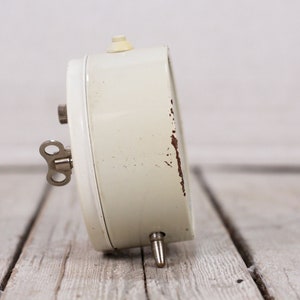 May include: A white wind-up alarm clock with a silver key. The clock has a white face and a silver knob on the top.