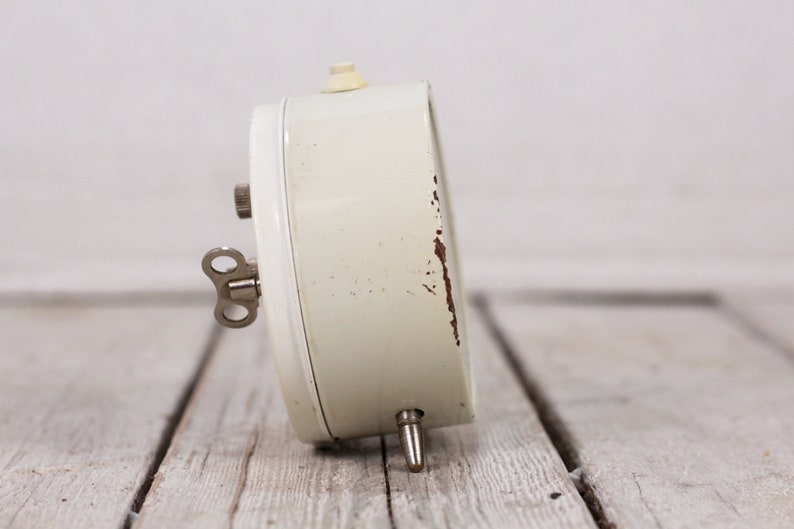 May include: A white wind-up alarm clock with a silver key. The clock has a white face and a silver knob on the top.
