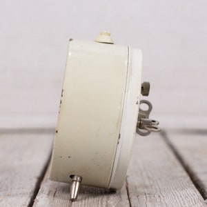 May include: A white vintage wind-up alarm clock with a key on the side. The clock is on a light wood surface.
