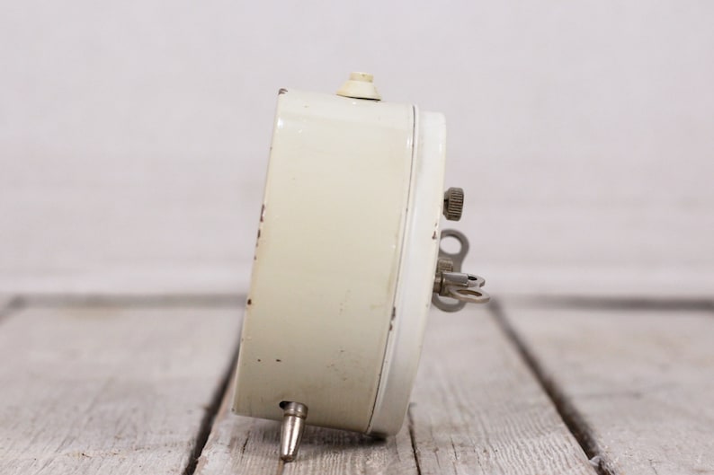 May include: A white vintage wind-up alarm clock with a key on the side. The clock is on a light wood surface.