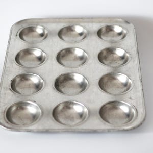 Pode incluir: Uma forma de muffin retangular e usada com doze cavidades redondas. A forma de metal tem um acabamento prateado e mostra sinais de desgaste. A assadeira é projetada para fazer muffins ou cupcakes.