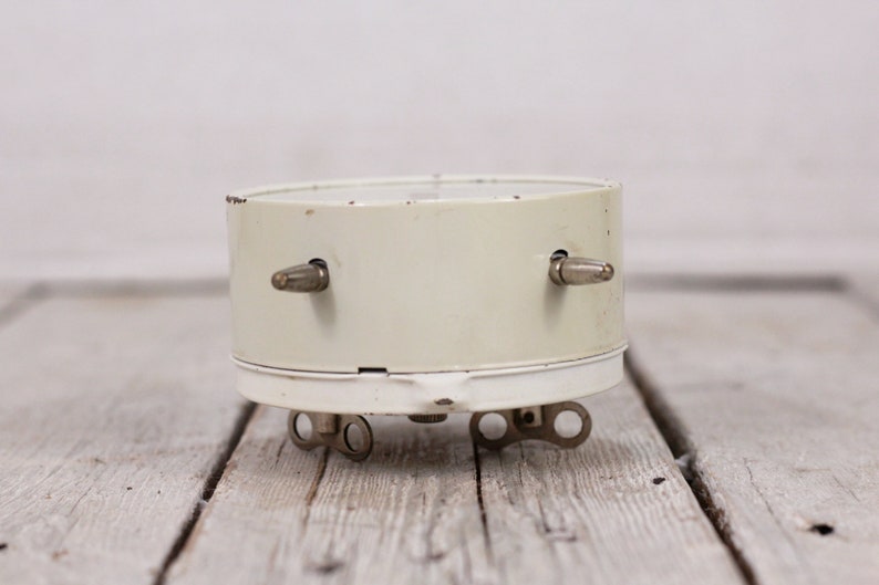 May include: A white vintage alarm clock with silver knobs on the side. The clock is sitting on a light wood surface.