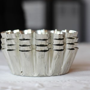 May include: A stack of four silver-colored metal baking molds with a fluted, scalloped edge design. Each mold features a circular base and vertical ridges along the sides, creating a consistent, repeating pattern throughout the nested metal set.