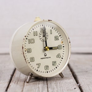 May include: White vintage alarm clock with black numbers and hands. The clock face reads "MEPRA-POLITIK".