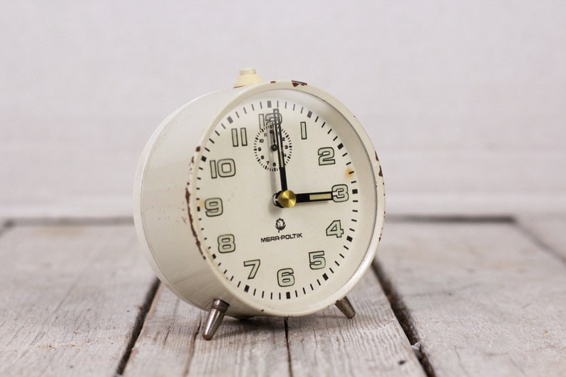 May include: White vintage alarm clock with black numbers and hands. The clock face reads "MEPRA-POLITIK".