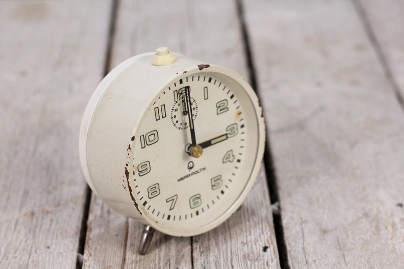 May include: White vintage alarm clock with black hands and numbers. The clock face has the text "MEWA POLTK" printed on it.